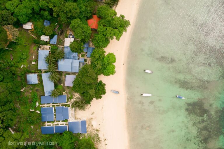 Pandangan udara pantai berpasir putih Pulau Tenggol dengan chalet beratap biru di tepi hutan dan bot berlabuh di air jernih