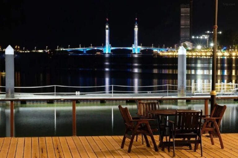 Dek kayu Duyong Marina & Resort pada waktu malam dengan meja dan kerusi, menghadap sungai dan jambatan berlampu di seberang
