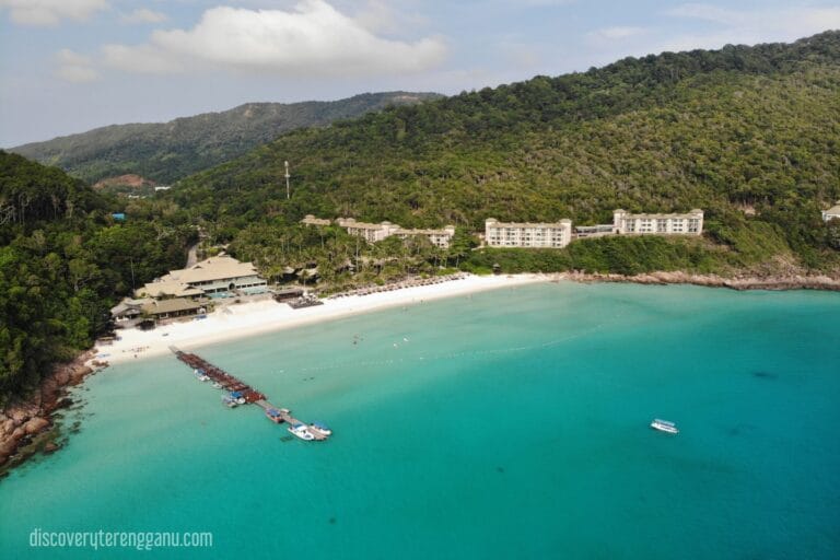 Pemandangan udara teluk di Pulau Redang dengan pantai pasir putih, jeti kayu, beberapa bot dan bangunan penginapan di lereng bukit berhutan