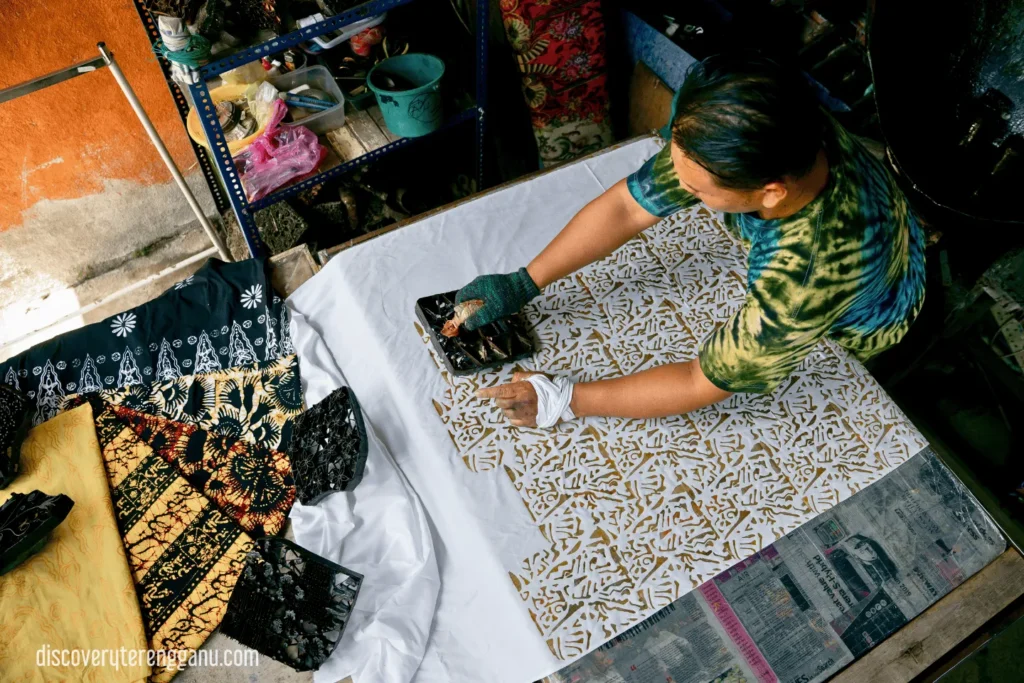 Tukang batik menekan blok logam bercorak ke atas kain putih yang terbentang di meja, meninggalkan corak berwarna emas; blok-blok lain dan kain siap di tepi meja