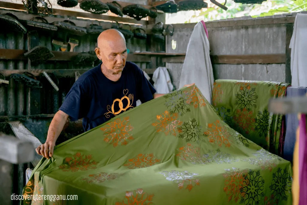 Tukang batik lelaki membentangkan kain hijau bercorak bunga terap, dengan deretan blok batik tersangkut di dinding bengkel di belakangnya