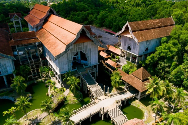 Pandangan udara Kompleks Muzium Negeri Terengganu di Bukit Losong dengan bangunan bertiang berbumbung tradisional, jambatan pejalan kaki dan pokok kelapa