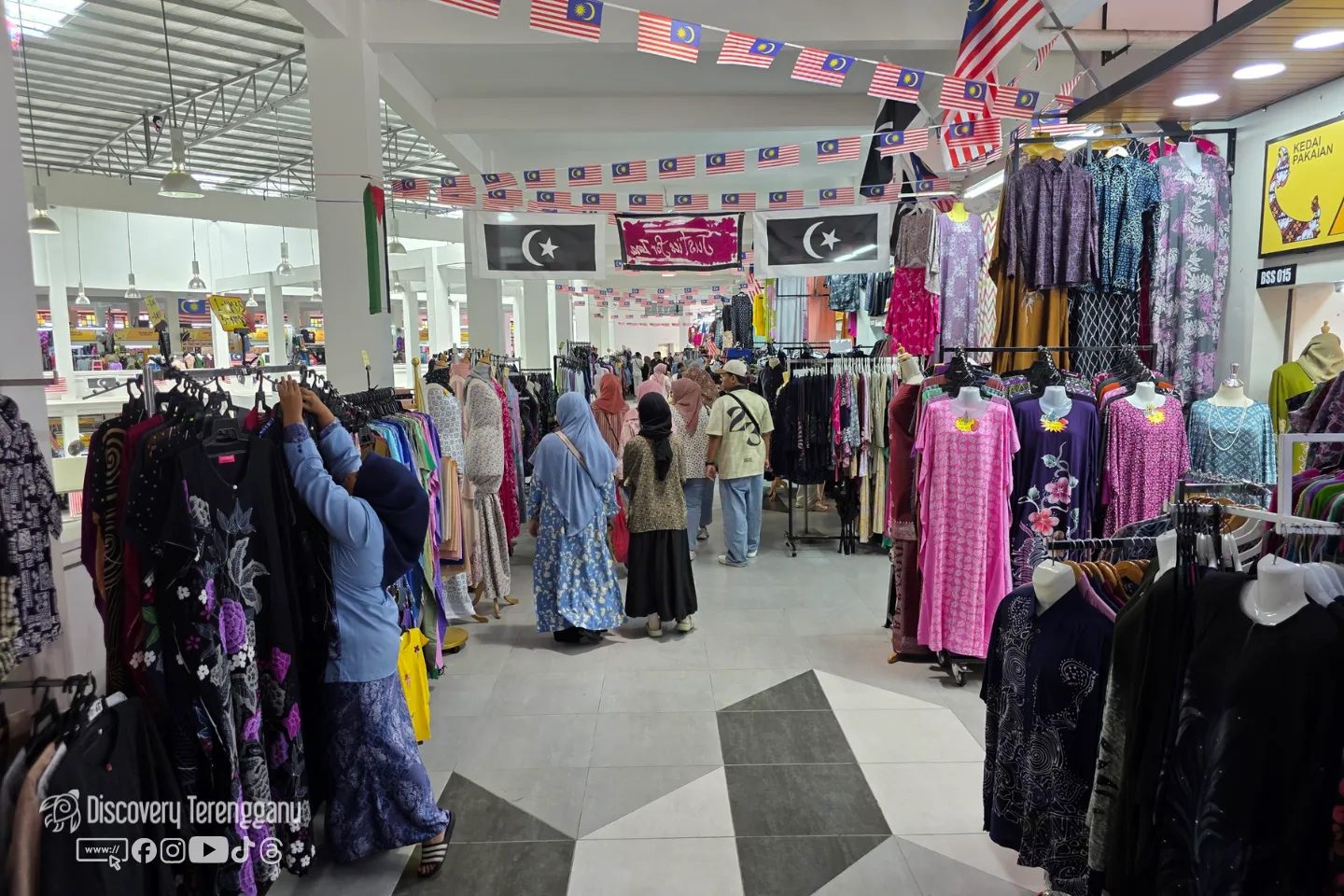 Lorong kedai pakaian di Pasar Kedai Payang dengan rak baju di kiri dan kanan serta bendera Malaysia dan Terengganu tergantung di siling