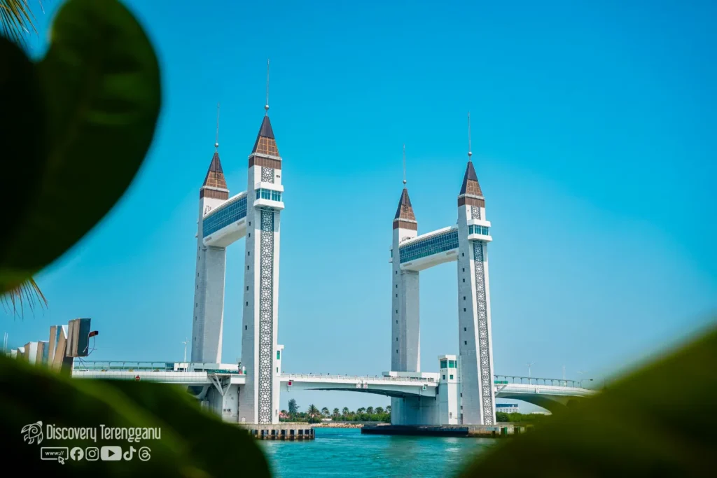 Jambatan Angkat Kuala Terengganu dengan empat menara putih berbumbung coklat di bawah langit biru, dilihat dari celah daun