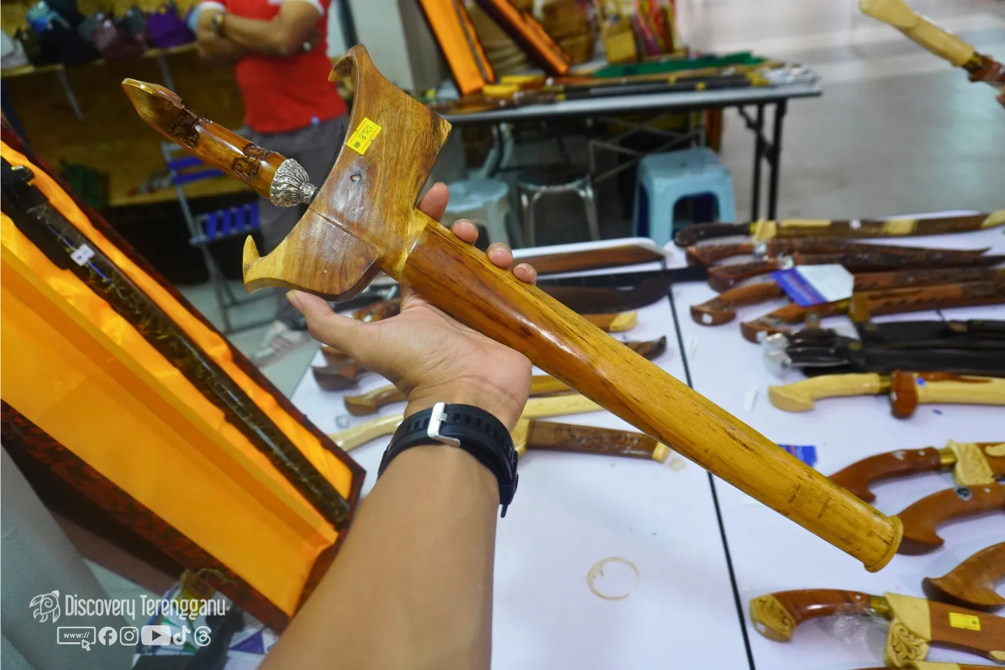 Tangan memegang sebilah keris bersarung kayu, dengan meja penuh keris dan sarung pelbagai ukiran di belakang