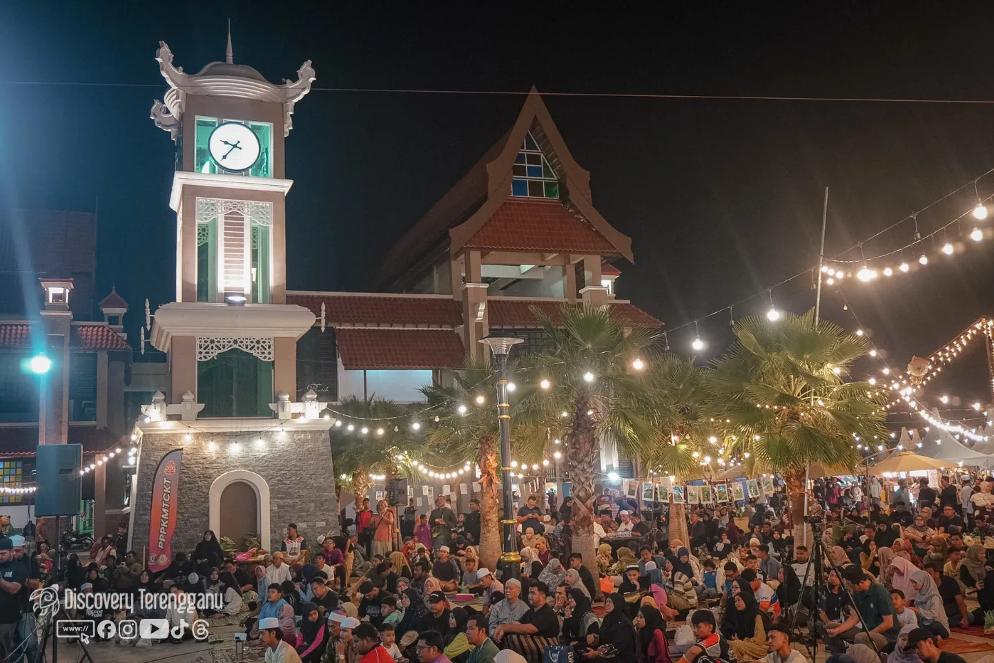 Orang ramai duduk di atas tikar pada waktu malam di perkarangan Pasar Kedai Payang dengan lampu berjuntai dan menara jam yang bercahaya